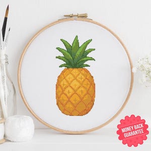 Pode incluir: Bordado em ponto cruz de um abacaxi num bastidor de madeira. O abacaxi é amarelo e laranja com folhas verdes. O bordado está em tecido branco. Um autocolante vermelho "Garantia de reembolso" é visível.