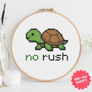 Può includere: Ricamo a punto croce in un telaio di legno con una tartaruga verde e marrone pixelata. Le parole "no rush" sono ricamate sotto in nero e verde. Il disegno è su tessuto bianco.