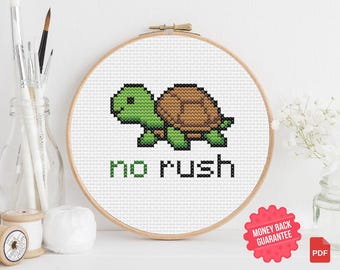 May include: Cross-stitch embroidery in a wooden hoop featuring a pixelated green and brown turtle. The words "no rush" are stitched below the turtle in black and green. The design is on white fabric.