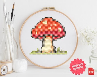 May include: A cross-stitch pattern of a red and white mushroom with a brown stem and green grass. The pattern is on a white background and is framed in a wooden hoop.