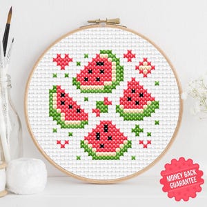 Pode incluir: Um bastidor de bordado em ponto cruz com um desenho de melancia. O desenho inclui quatro fatias de melancia em vermelho, verde e preto, com detalhes adicionais em ponto cruz verde e vermelho. O bastidor é exibido em uma superfície branca.