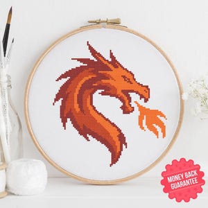 Può includere: Un disegno a punto croce di una testa di drago nei toni dell'arancione e del rosso, incorniciato in un telaio da ricamo in legno. Il drago sputa fuoco. L'immagine include un adesivo "Soddisfatti o rimborsati" e un'icona PDF.