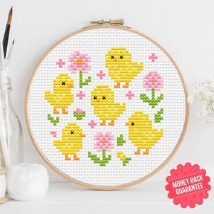 Può includere: Un telaio da ricamo a punto croce con un disegno di pulcini gialli e fiori rosa su sfondo bianco. Il ricamo è inserito in un telaio di legno. L'immagine include un adesivo "Soddisfatti o rimborsati" e un'icona PDF.