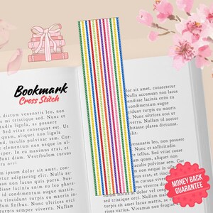Könnte beinhalten: Ein farbenfrohes Kreuzstich-Lesezeichen mit vertikalen Streifen in verschiedenen Farben, das in einem offenen Buch liegt. Das Lesezeichen zeigt den Text "Bookmark Cross Stitch". Rosa Blüten und Blütenblätter sind verstreut.
