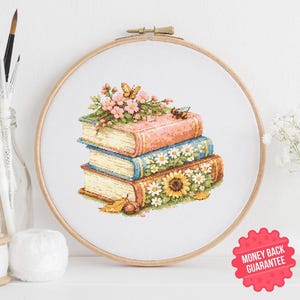 May include: Cross-stitch embroidery of three stacked books adorned with flowers, a butterfly, and a bee. The books have pink, blue, and brown covers. The embroidery is set in a wooden hoop, with a white background.