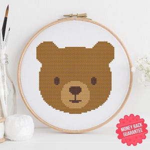 May include: A cross-stitch embroidery hoop featuring a brown bear face design. The bear's head is centered on white fabric within a wooden hoop. The design includes a nose, eyes, and mouth. The image also includes a "Money Back Guarantee" sticker.