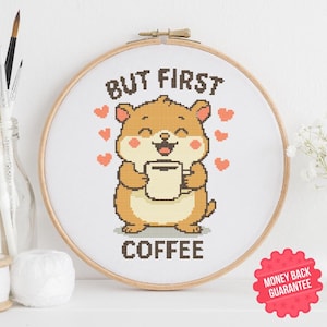 May include: A cross-stitch embroidery hoop with a cartoon hamster holding a coffee cup. The text "BUT FIRST COFFEE" is stitched below the hamster. The design includes small red hearts and is set against a white background.