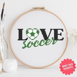 May include: A white embroidery hoop with black and green text that reads "LOVE soccer" with a green and white soccer ball in the shape of a heart.