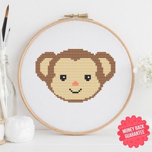 May include: A cross-stitch monkey face displayed in a wooden embroidery hoop. The monkey features a light brown face, dark brown ears and head, and a peach nose. The background is white. The image includes a "Money Back Guarantee" sticker.