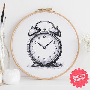 May include: A black and white cross-stitch design of an alarm clock, set in a wooden embroidery hoop. The clock face shows the time with hands pointing to the numbers. The design is displayed on a white surface.