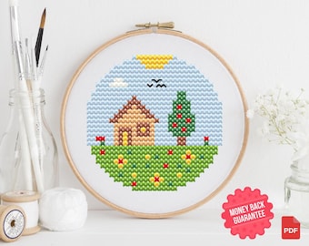 May include: A cross-stitch embroidery hoop featuring a landscape scene. The design includes a brown house, a green tree with red accents, and a grassy field with flowers. The sky is blue with a yellow sun and black birds. The hoop is set against a white background.