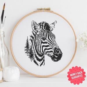 Op de afbeelding: Een zwart-wit kruissteekborduurwerk van een zebrahoofd, gepresenteerd in een houten borduurring. Het gezicht van de zebra is gedetailleerd met strepen. De achtergrond is wit. Een rode sticker "Geld terug garantie" is zichtbaar.