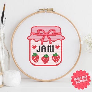 Op de afbeelding: Een kruissteekontwerp van een roze jam pot met het woord "JAM" en drie aardbeien. Het ontwerp is ingelijst in een houten borduurring. De afbeelding bevat een "Geld terug garantie" sticker en een PDF-icoon.