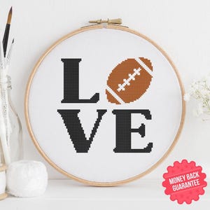 May include: A cross-stitch embroidery hoop featuring the word "LOVE" with a football replacing the "O". The letters are black, and the football is brown with white stitching. The hoop is set against a white background.