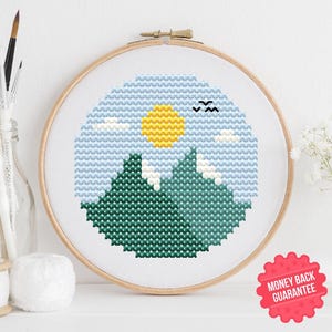 May include: A cross-stitch embroidery hoop featuring a landscape design. The artwork depicts green mountains, a yellow sun, blue sky, and small black birds. The design is set against a white background, with a wooden hoop.