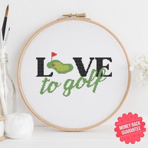 Puede incluir: Un aro de bordado blanco con un diseño de punto de cruz que dice "Love to golf" con un campo de golf verde y una bandera en el medio de la palabra "Love".