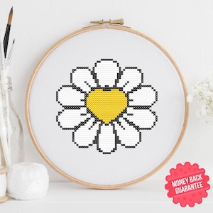 May include: A cross-stitch embroidery hoop with a daisy design. The daisy has a yellow heart-shaped centre and white petals, set in a wooden hoop. A red sticker with "MONEY BACK GUARANTEE" is in the lower right corner.