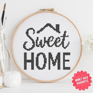 May include: A cross-stitch embroidery hoop with the words "Sweet Home" in black on white fabric. The design includes a house silhouette above the word "Sweet". The hoop is displayed on a white surface with art supplies and flowers in the background.