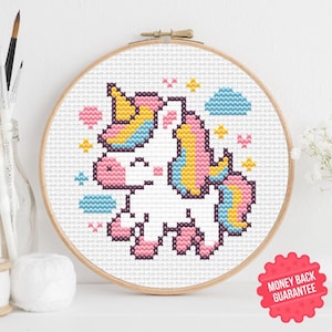 Puede incluir: Patrón de punto de cruz de un unicornio con cuerno amarillo, melena y cola rosas y detalles azules. El diseño está enmarcado en un aro de madera, sobre un fondo blanco. Incluye una pegatina de "Garantía de devolución de dinero".