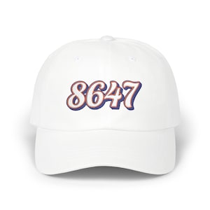 May include: A white baseball cap with the number "8647" embroidered in a stylized font. The numbers are white with a blue outline and a reddish-orange shadow, creating a 3D effect. The cap has a curved brim and a button on top.
