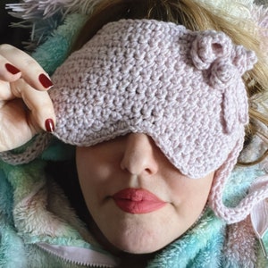 May include: A close-up of a person wearing a pink crocheted sleep mask with a flower detail. The mask is covering the person's eyes. The person is wearing a blue and green fuzzy hooded jacket.