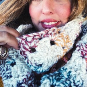 May include: A chunky, handmade crochet cowl scarf in a mix of white, teal, burgundy, and yellow. The scarf is made with a thick yarn, creating a textured, cozy look. The cowl is designed to be worn around the neck for warmth and style.