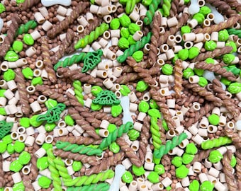 May include: A mix of brown, green, and white pasta shapes, including twists, rings, and small green leaves. The pasta is in a bowl and appears to be ready to be used for a craft project.