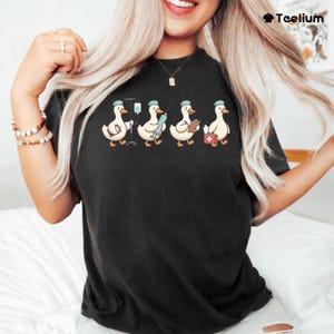 以下が含まれることがあります： 医療従事者の格好をした漫画のアヒルのデザインが施された黒いTシャツ。アヒルは青い帽子をかぶり、点滴、注射器、クリップボード、救急箱などの医療品を運んでいます。Tシャツはクルーネックです。