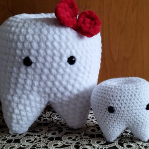 May include: Two white crocheted plush teeth, one larger than the other. The larger tooth has a red bow on top. Both teeth have black eyes.
