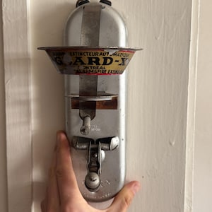 May include: A vintage silver-toned metal Guard-X automatic fire extinguisher device featuring a conical top section with red and yellow text, a central vertical mounting bracket, and a spring-loaded mechanical release lever at the bottom.