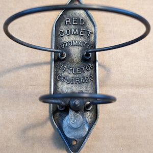 Puede incluir: Un soporte metálico antiguo para extintor con el texto "RED COMET AUTOMATIC LITTLETON COLORADO". El soporte es de color metálico oscuro con dos soportes de alambre circulares.