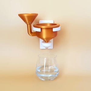 Könnte beinhalten: Ein orange-weißes, an der Wand montiertes Wasserspendersystem mit Trichterdesign. Darunter steht ein klares Wasserglas. Der Spender besteht aus Kunststoff und hat eine moderne, minimalistische Ästhetik.