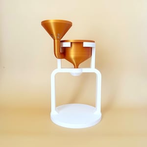 Può includere: Una caffettiera stampata in 3D in bianco e bronzo. Il design presenta un imbuto, una camera di infusione e una base. I componenti in bronzo sono collegati da un telaio bianco. La caffettiera è progettata per essere posizionata su una superficie piana.