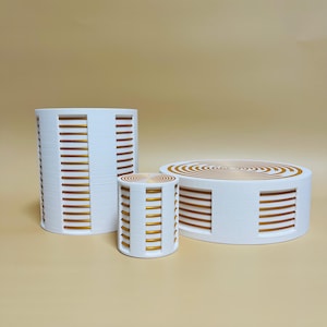 Op de afbeelding: Drie witte cilindrische displaystandaards met gouden accenten. De grootste standaard is hoog en smal, de middelste standaard is kort en breed, en de kleinste standaard is kort en smal.