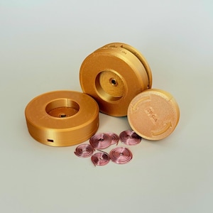 Può includere: Una collezione di bobine color rame e un disco rotondo con la scritta "20 GA" e frecce. Sono presenti anche diverse piccole bobine di filo rosso. Gli oggetti sono disposti su una superficie grigio chiaro.