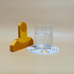 Könnte beinhalten: Ein weißer Untersetzer mit goldenen Akzenten und einem Glas Wasser darauf. Der Untersetzer ist aus einem 3D-gedruckten Material gefertigt und hat ein kreisförmiges Design. Ein goldfarbenes 3D-gedrucktes Objekt befindet sich im Hintergrund.