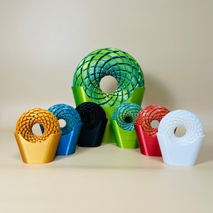 Può includere: Una collezione di sculture astratte stampate in 3D dai colori vivaci. Le sculture presentano un design circolare intrecciato con un foro centrale. I colori includono oro, blu, nero, verde, rosso e bianco. La scultura verde più grande è in un supporto verde.