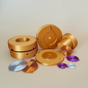 Può includere: Una collezione di componenti meccanici circolari color oro e molle. I componenti includono dischi, cilindri e una pila di anelli di varie dimensioni. Sono presenti anche diverse molle a spirale colorate in argento, rame e viola.