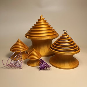 Può includere: Quattro oggetti a forma di spirale di colore dorato, stampati in 3D. L'oggetto più grande ha una base larga e una punta appuntita. Gli altri tre oggetti sono più piccoli e hanno una forma più arrotondata. L'oggetto più piccolo è una spirale viola.