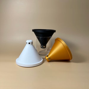 Op de afbeelding: Drie keramische koffiefilters voor gieten in verschillende kleuren: wit, zwart en goud. De filters zijn kegelvormig en hebben een klein gaatje aan de onderkant.