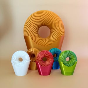 Può includere: Un set di cinque oggetti a forma di ciambella stampati in 3D e colorati. La ciambella più grande è dorata e le ciambelle più piccole sono bianche, rosse, blu e verdi. Ogni ciambella è posizionata in un supporto dello stesso colore.