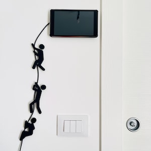 Puede incluir: Tableta negra montada en una pared blanca con un diseño decorativo de figuras negras escalando. El diseño presenta tres figuras de palo subiendo por una cuerda, añadiendo un toque único a la decoración de la pared. La tableta está conectada a la figura.