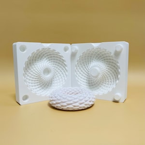 Może przedstawiać: Dwie białe, otwarte połówki formy z zakręconym, fakturowanym wzorem. Biały, wydrukowany w 3D pierścień z podobnym zakręconym wzorem znajduje się przed formami.