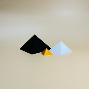 Può includere: Tre piramidi di carta di diverse dimensioni. La piramide più grande è nera, la piramide di mezzo è dorata e la piramide più piccola è blu chiaro.