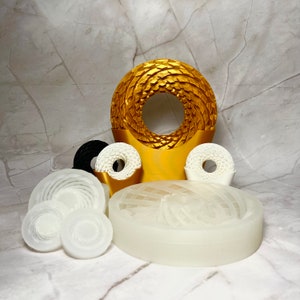 May include: A set of white and black silicone moulds for making circular cakes. The moulds feature a variety of geometric patterns, including a spiral, a honeycomb, and a circle. The moulds are arranged on a white marble surface.