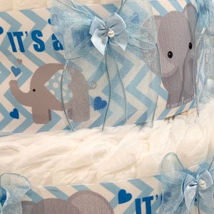 Pode incluir: Um bolo de fraldas azul e branco com uma fita e um la&ccedil;o azul. O bolo &eacute; decorado com elefantes cinzentos e o texto "&Eacute; um menino".
