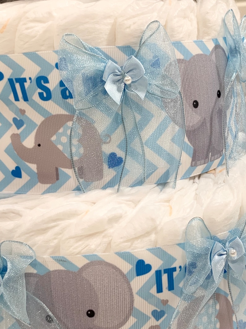 Pode incluir: Um bolo de fraldas azul e branco com uma fita e um la&ccedil;o azul. O bolo &eacute; decorado com elefantes cinzentos e o texto "&Eacute; um menino".