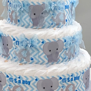 Pode incluir: Um bolo de fraldas de tr&ecirc;s camadas decorado com papel estampado em chevron azul e branco, figuras de elefantes cinzentos e la&ccedil;os azuis. O bolo &eacute; coroado com um elefante de pel&uacute;cia cinza. A camada inferior tem o texto "It's a boy" impresso nela.