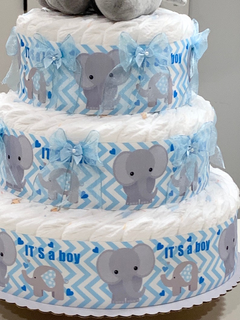 Pode incluir: Um bolo de fraldas de tr&ecirc;s camadas decorado com papel estampado em chevron azul e branco, figuras de elefantes cinzentos e la&ccedil;os azuis. O bolo &eacute; coroado com um elefante de pel&uacute;cia cinza. A camada inferior tem o texto "It's a boy" impresso nela.