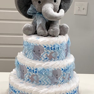Pode incluir: Um bolo de fraldas de tr&ecirc;s camadas decorado com tecido estampado em chevron azul e branco e aplica&ccedil;&otilde;es de elefante cinza. A camada superior apresenta um elefante de pel&uacute;cia cinza com uma fita azul.